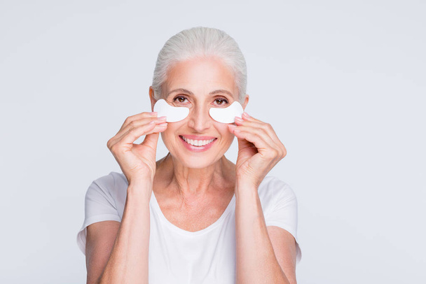 Close up photo of sweet grandmother holding cotton pad under eyes applying lotion smiling wearing stylish trendy t-shirt isolated over white background - 写真・画像