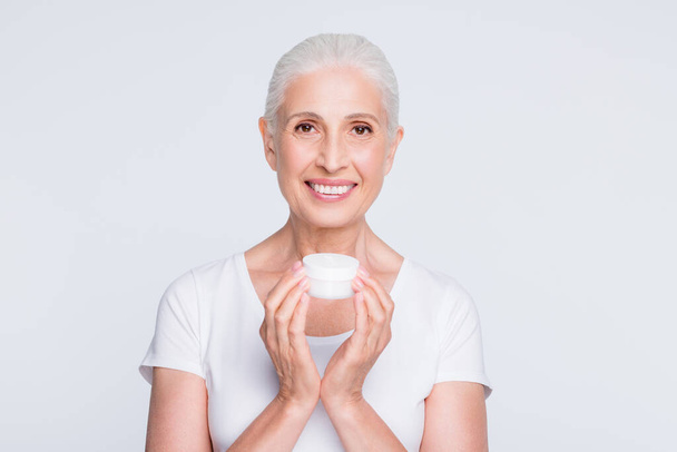 Portrait of lovely grandmother holding jar with nourishing procedure looking at camera wearing stylish trendy t-shirt isolated over white background - Фото, зображення