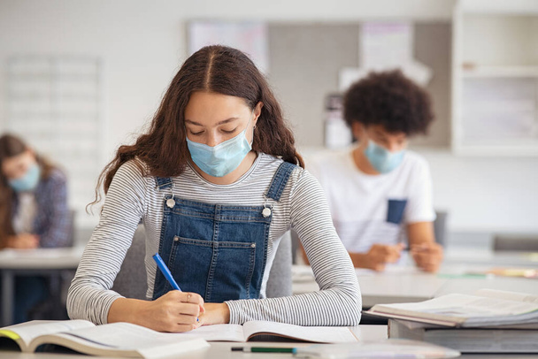 High school student taking notes from book while wearing face mask due to coronavirus emergency. Young woman sitting in class with their classmates and wearing surgical mask due to Covid-19 pandemic. Focused girl studying in classroom. - Foto, imagen