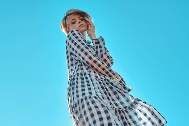 Menina elegante bonito em um vestido xadrez posando contra o céu azul. - Foto, Imagem