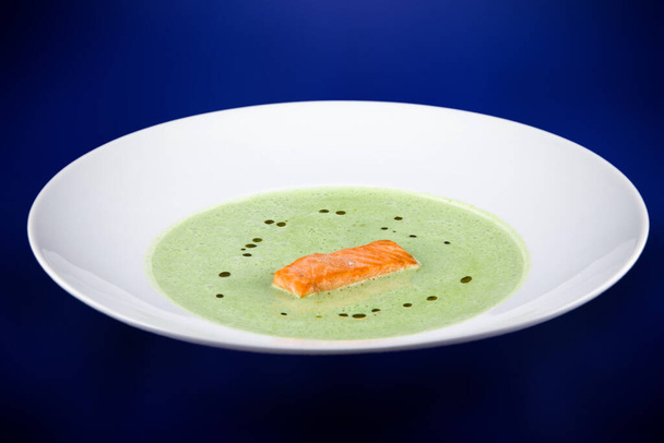 Green spinach puree soup with salmon slices. Flat lay top view on a white plate on a blue background. - Photo, Image