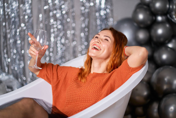 The beautiful red-haired girl rejoices sitting dressed in the bath. against the background of tinsel and balloons. Success concept. - Foto, afbeelding