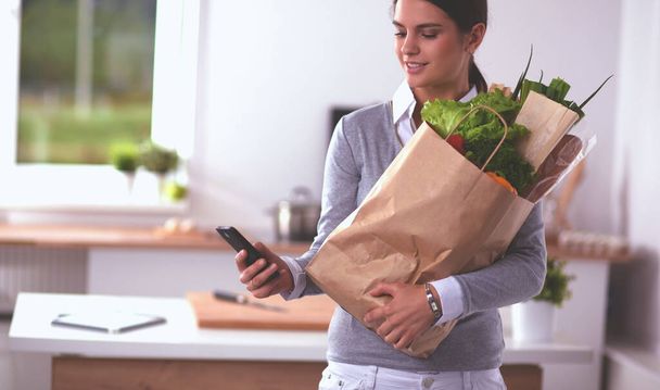 Smiling woman with mobile phone holding shopping bag in kitchen - Foto, Imagem