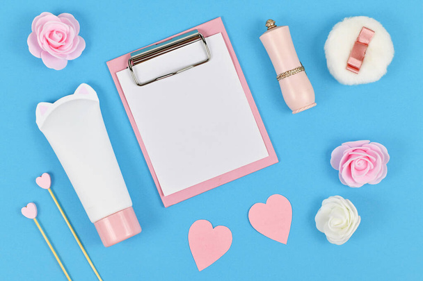 Flat lay with blank clipboard and rose flowers, cream tube in shape of cat, paper hearts and powder puff on blue background  - Foto, Imagen
