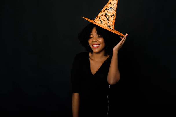 ハロウィンオレンジの帽子と黒のドレスを身に着けているアフロヘアカットを持つ楽しい若い黒人女性の水平ショット。黒を背景にしたスタジオポートレート - 写真・画像