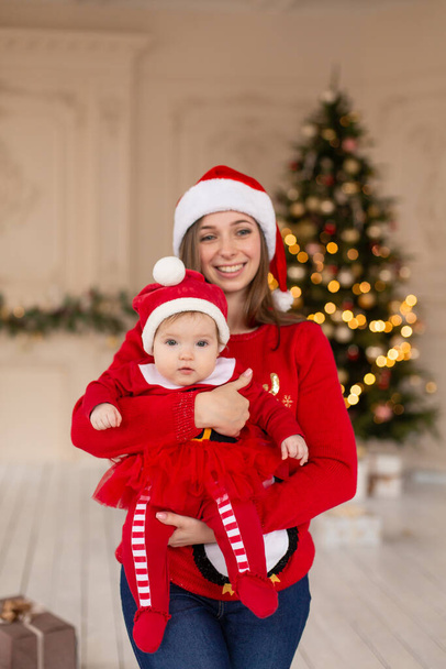 Happy mother hug little daughter in red Santas costume and hat, near Christmas tree. - Фото, изображение