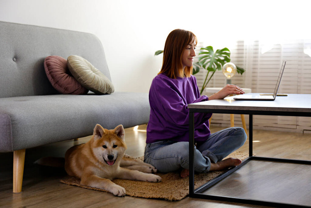 Portrait of a woman sitting on the floor, working on a laptop at home with her nine months old japanese akita inu pup. Happy female and funny big breed dog relaxing at home. Close up, copy space. - Φωτογραφία, εικόνα