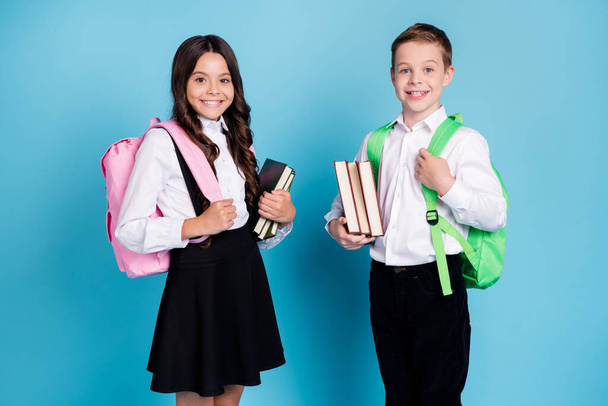 Photo of two small girl boy schoolchildren brother sister classmates hold books walk library study year finishing wear backpack white shirt black pants dress isolated blue color background - Fotó, kép