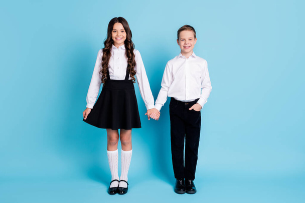 Full length photo of two girl boy schoolchildren best friends shiny smiling hold hands first school day wear uniform white shirt black pants dress shoes isolated blue color background - Φωτογραφία, εικόνα