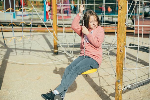 Χαριτωμένο καυκάσιος κορίτσι 7-8 ετών σε ένα ροζ φούτερ και γκρι φόρμα στην παιδική χαρά στο πάρκο της πόλης. Υπαίθριες δραστηριότητες, τρόπος ζωής, ψυχαγωγία των παιδιών - Φωτογραφία, εικόνα