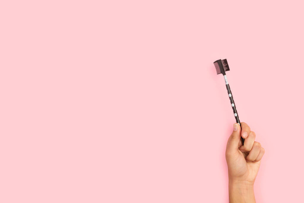 Mano de mujer sosteniendo un peine de cejas sobre un fondo rosa con espacio para copiar - Foto, imagen
