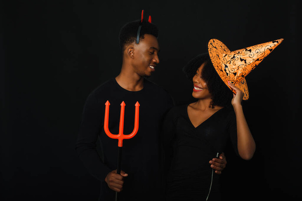 Una pareja afectuosa pareja afroamericana celebra Halloween en el estudio con disfraces sobre un fondo negro. - Foto, imagen