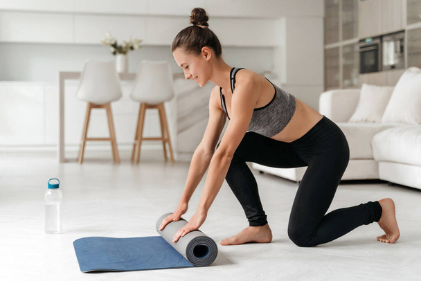 Adaptez jeune femme déroulant tapis de yoga et se préparant pour l'entraînement de remise en forme ou cours de yoga à la maison. Pilates roulants féminins sportifs installations sportives après l'entraînement. Concept de mode de vie sain. Fond de pièce - Photo, image