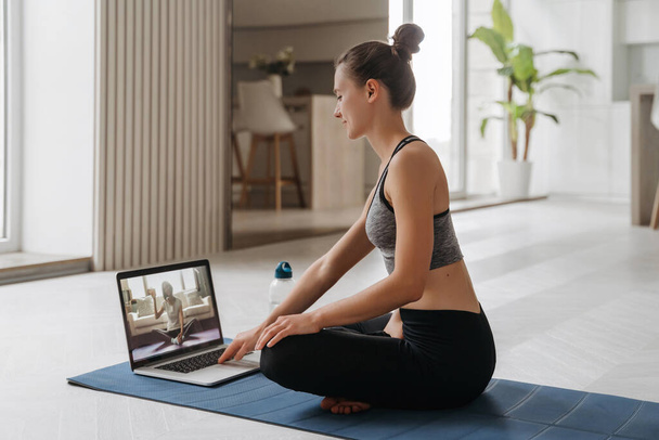 Вчитель йоги проводить вдома віртуальний клас йоги на відеоконференції. Прекрасна жінка, яка практикує йогу онлайн у своїй вітальні з ноутбуком. Концепція домашнього фітнесу та тренування. Онлайн-навчання - Фото, зображення