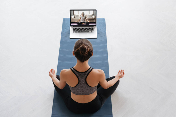 Top vue jeune femme sportive à l'aide d'un ordinateur portable pour la formation hatha yoga avec instructeur virtuel, faire Sukhasana, se détendre et respirer facilement. Fitness en ligne, entraînement à la maison. Bien-être, concept de mode de vie actif - Photo, image
