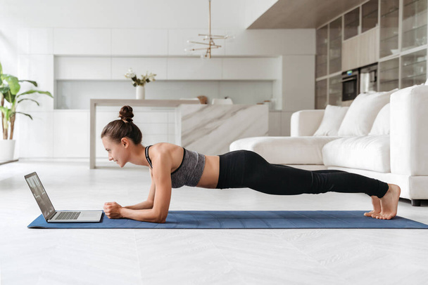 Beautiful young woman in sports uniform doing plank exercise in living room at home, watching videos on laptop computer and repeating online instructions. Online workout. Copy space. Full length side - 写真・画像