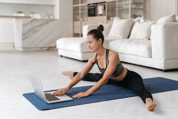 Fit young woman doing stretching on mat at home in her living room, using laptop for watching virtual tutorial, repeating exercising, has online fitness class. Wellness, healthy lifestyle concept  - Фото, изображение