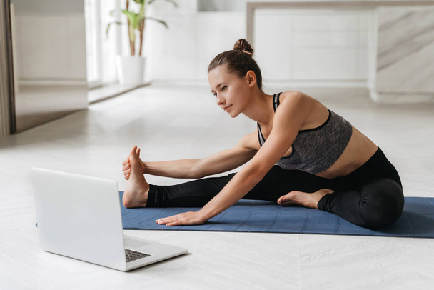 Young sporty woman doing yoga exercises while sitting on mat in her room. Stretching instructor conducts virtual class on video conference using laptop. Home interior background. Stretching, wellness - 写真・画像