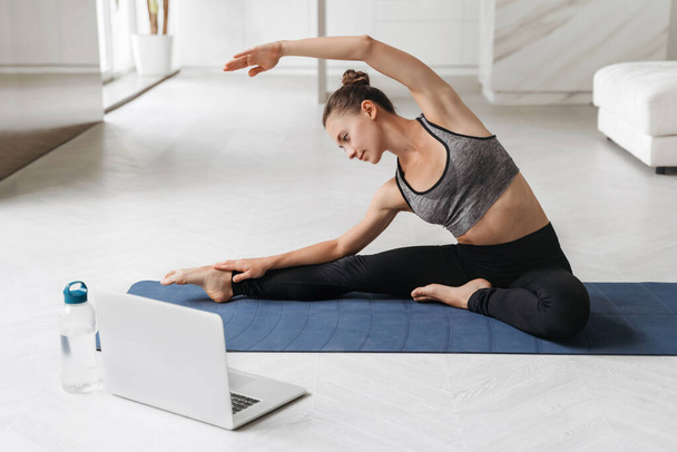 Young sporty woman exercising while sitting on yoga mat in her apartment. Active fit female doing stretching exercise for arms and back in living room at home. Wellness, activity, healthy lifestyle - Photo, Image