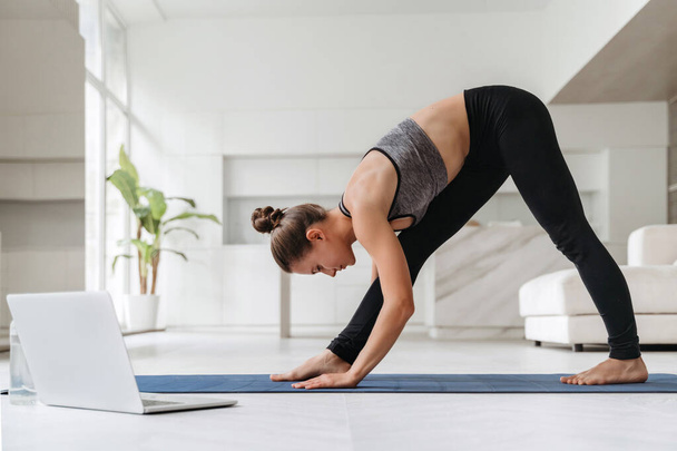 Verbazingwekkende jonge mooie brunette fitness vrouw doen stretching oefeningen binnen in de buurt van laptop computer. Fit vrouw doet yoga, fitness, workout thuis. Wellness en gezonde levensstijl concept - Foto, afbeelding