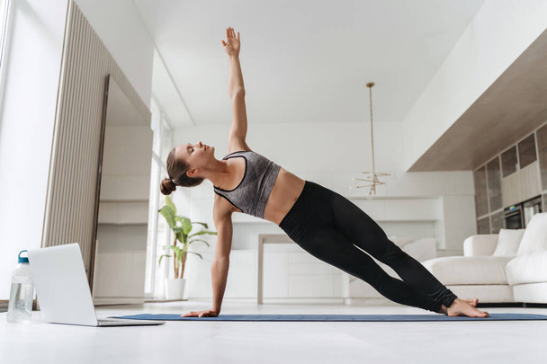 Hübsches Mädchen mit Leggings und kurzem Oberteil, das in der Seitenplanke auf einer Hand zu Hause steht, trainiert Körperkern und Gleichgewicht und stärkt die Muskeln. Attraktive Frau praktiziert Yoga in ihrem Zimmer mit Laptop - Foto, Bild