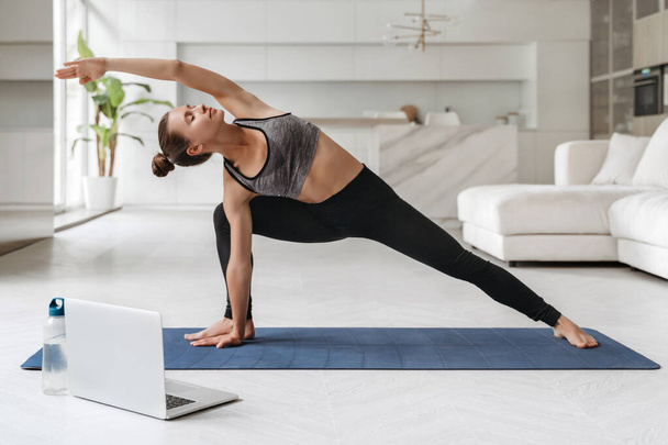 Mujer atractiva joven que practica yoga y estira el cuerpo en casa usando el portátil para clases en línea o tutoriales virtuales. Chica de pie en Utthita parsvakonasana ejercicio, haciendo Side Angle pose. Bienestar - Foto, Imagen