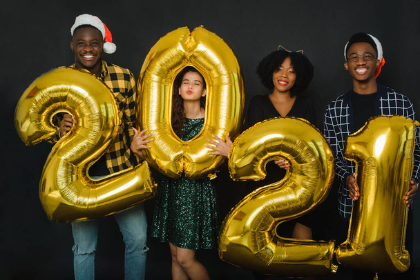 Un grupo de jóvenes amigos de diferentes razas están celebrando la Navidad en el estudio. Jóvenes diversos sostienen números de oro 2021 y bengalas en el fondo negro. - Foto, imagen