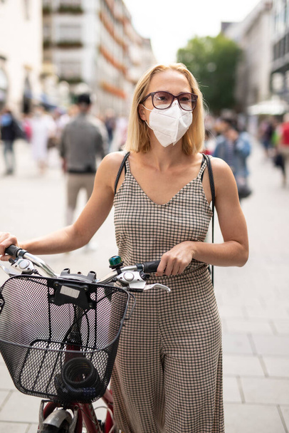 Woman walking by her bicycle on pedestrian city street wearing medical face mask in public to prevent spreading of corona virus. New normal during covid epidemic. - Photo, Image