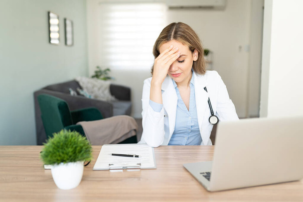 Female doctor touching her forehead and looking stressed and overwhelmed while doing some paperwork at home - Foto, Imagen