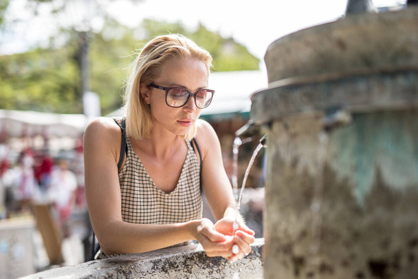 Διψασμένη νεαρή λευκή γυναίκα πίνει νερό από το σιντριβάνι της πόλης σε μια ζεστή καλοκαιρινή μέρα - Φωτογραφία, εικόνα
