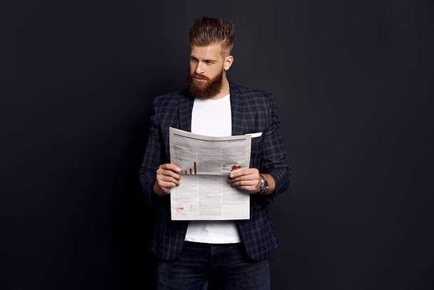 Γενειοφόρος έξυπνος όμορφος συγκεντρωμένος αρχηγός ντυμένος με σακάκι και πουκάμισο διαβάζοντας τα τελευταία νέα από εφημερίδα απομονωμένη στα μαύρα. - Φωτογραφία, εικόνα