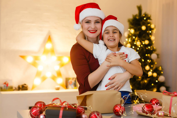 Vidám csinos anya és imádnivaló kislánya élvezi szépségét kézzel készített karácsonyi dekoráció, miközben ül az asztalnál, belső hangulatos nappali a háttérben - Fotó, kép