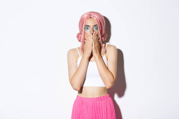 Portrait of shocked woman in pink party wig looking ambushed, gasping and covering mouth with hands, seeing something shocking or scary, wearing halloween costume - 写真・画像
