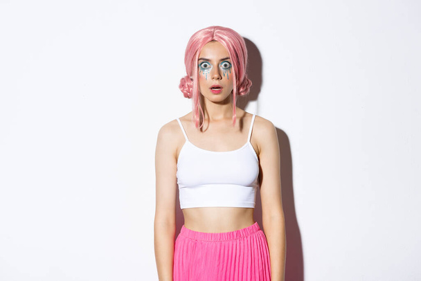 Πορτρέτο του λαμπερό κορίτσι κόμμα σε ροζ περούκα και φωτεινό μακιγιάζ, πτώση σαγόνι και αναζητούν τρόμαξε στην κάμερα, κοιτάζω κάτι εντυπωσιακό, στέκεται πάνω από το λευκό φόντο - Φωτογραφία, εικόνα
