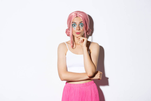 Захватывающая милая тусовщица в розовом парике и хэллоуинском макияже, с интересом смотрит в камеру, слушает что-то, стоит на белом фоне - Фото, изображение