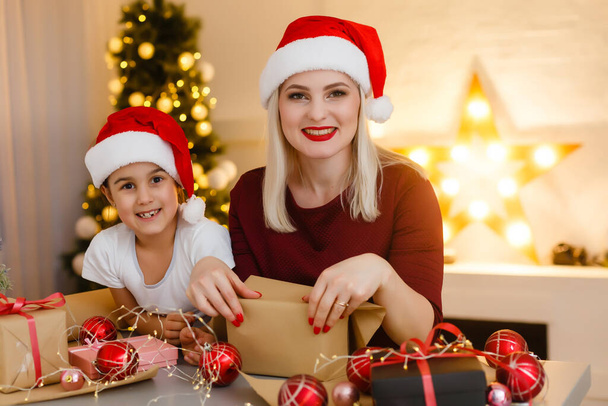 Iloinen kaunis äiti ja hänen suloinen pieni tyttärensä nauttivat kauneudesta käsintehty joulukoristeen istuessaan pöydällä, sisätilojen viihtyisä olohuone taustalla - Valokuva, kuva