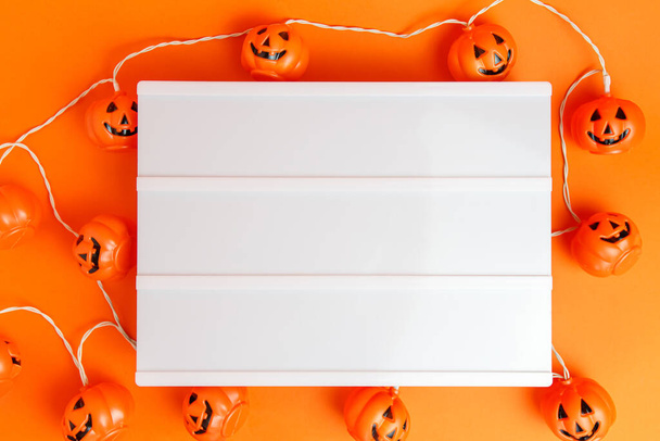 Белая доска с маленькой страшной тыквой на оранжевом фоне. Хэллоуин. - Фото, изображение