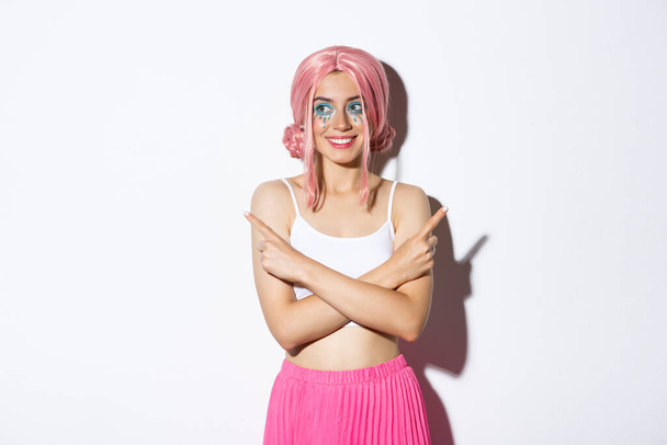 Belle fille de fête souriante en costume d'Halloween et perruque rose, pointant du doigt latéralement mais regardant à gauche, faisant un choix, debout sur fond blanc - Photo, image