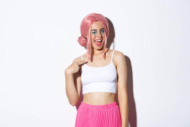 Image d'une jolie fille effrontée en perruque rose et tenue de fête, clignant des yeux et se pointant du doigt, portant un costume d'Halloween, debout sur fond blanc - Photo, image