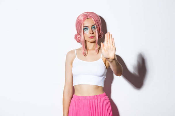 Bild eines ernsthaften weiblichen Modells in rosa Perücke und Halloween-Kostüm mit Stoppschild, ausgestreckte Hand, um etwas Schlechtes abzulehnen oder zu verbieten, vor weißem Hintergrund stehend - Foto, Bild
