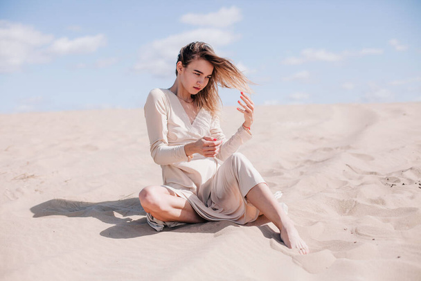 A young, slender girl in a beige dress poses in the wind in the desert - Photo, image