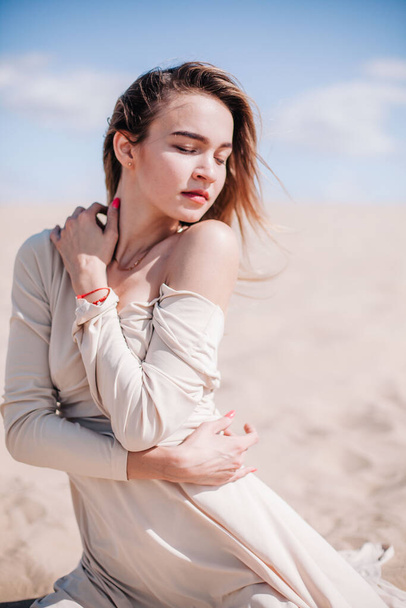 A young, slender girl in a beige dress poses in the wind in the desert - Foto, Imagen