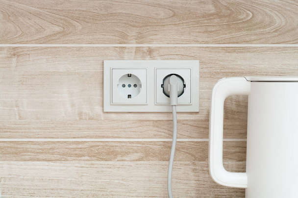 Chaleira elétrica branca conectada a uma tomada de energia em uma parede em uma cozinha - Foto, Imagem
