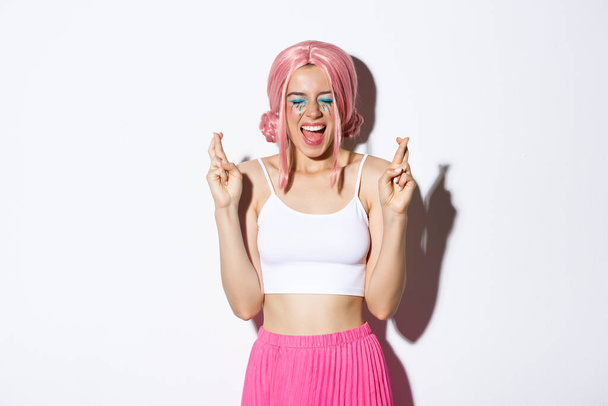 Wizerunek optymistycznej dziewczyny w różowej peruce, z makijażem halloween, zamkniętymi oczami i krzykiem podekscytowany podczas krzyżowania palców, aby życzenie, stojąc nad białym tle - Zdjęcie, obraz