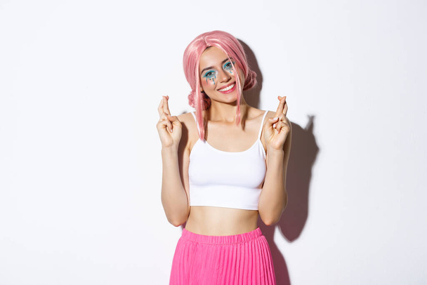 Portrait of lovely smiling woman in halloween costume, pink wig and bright makeup, looking hopeful at camera and making wish with fingers crossed - Photo, Image