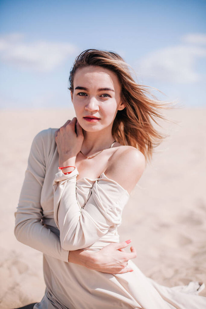 Egy fiatal, karcsú lány bézs ruhában pózol a szélben a sivatagban. - Fotó, kép