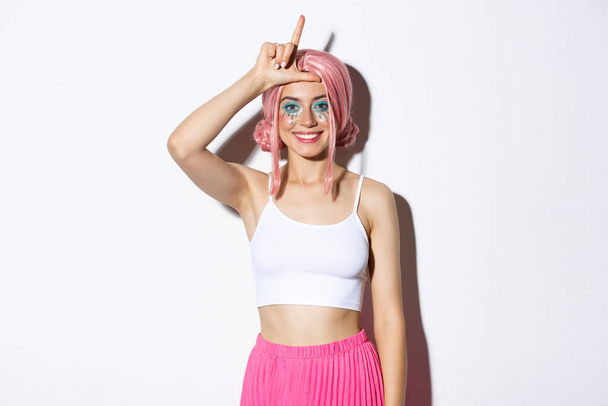Pretty smiling woman looking happy as winning, showing loser sign on forehead as mocking someone, standing over white background in halloween costume - Photo, Image
