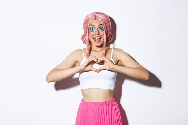 Obrázek vzrušené usměvavé dívky jako něco, ukazuje srdeční gesto a vypadá ohromeně, na sobě halloween kostým s jasným make-up a růžová paruka - Fotografie, Obrázek