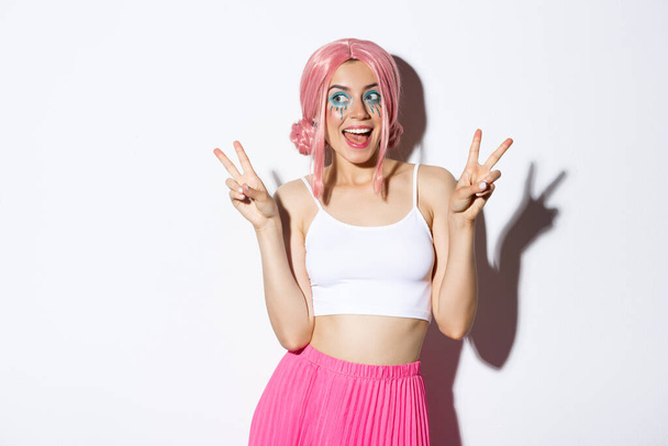 Retrato de menina atraente elegante em peruca rosa e traje de Halloween, mostrando sinais de paz e sorrindo feliz, de pé sobre fundo branco - Foto, Imagem