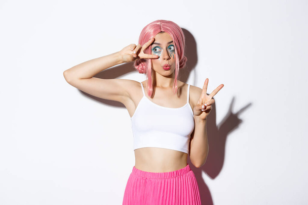 Image de silly beau modèle féminin avec perruque rose et maquillage d'Halloween, regardant à gauche surpris, faire des signes de paix, debout sur fond blanc - Photo, image
